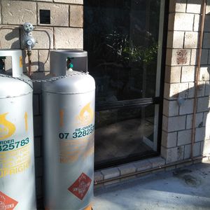 The image captures two large gas cylinders positioned beside a glass door. The cylinders are silver with warning labels and a visible re-order contact number. A gas meter is mounted on the wall, and some greenery is visible through the glass door, indicating an outdoor area.