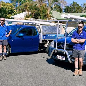 The image shows two utility vehicles parked on a residential street. One is a blue Toyota Hilux with a ladder rack, and the other is a blue Holden with similar fittings. Both vehicles are equipped for trades, featuring signage indicating their purpose and location. The background includes greenery and residential houses, suggesting a suburban environment.