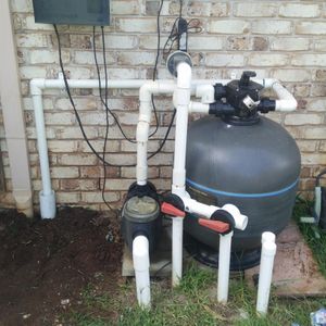 The image features a pool filtration system comprising a large cylindrical tank, several white PVC pipes, a control panel, and a clear pump. The setup is located outside, adjacent to a wooden fence, with some garden space visible in the foreground.