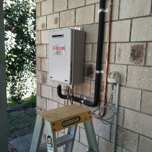 The image shows a Rinnai gas hot water system mounted on a wall. The unit is connected to pipes, with a ladder beneath it for installation or maintenance purposes. The surrounding wall is made of textured concrete blocks, and there are visible pipes leading from the unit.