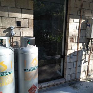 The image features two gas cylinders positioned next to a glass sliding door, leading into an interior space. Adjacent to the door is a gas meter and piping, indicating a setup for gas supply. The structure appears well-maintained, with brick walls and clear visibility of the surrounding area.