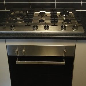 The image features a modern stainless steel gas cooktop with five burners and a sleek black oven below. The cooktop has various dials for adjusting heat and features a clean, contemporary design, set against a dark tiled backsplash.