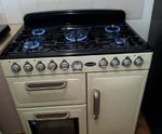 The image features a cream-coloured gas stove with a black grill top. It has four gas burners, each with visible blue flames, and knobs for adjusting heat levels. The stove also includes two oven compartments below, one with a glass door, suggesting it’s designed for both cooking and baking.