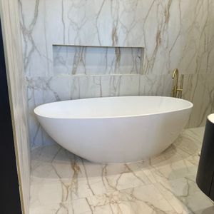 The image features a modern bathroom with a sleek, white freestanding bathtub positioned against a wall adorned with elegant marbled tiles. The interior design showcases a luxurious and contemporary style, with gold-finished fixtures complementing the marble's natural tones. The flooring continues the marble theme, enhancing the overall aesthetic of the space, which is well-lit and spacious.