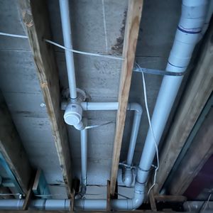 The image shows a ceiling with exposed pipes and wooden beams. Various plastic and metal plumbing pipes run across, indicating a partial construction or renovation phase in a building.