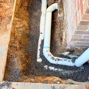 The image shows a trench dug alongside a brick wall, revealing two white PVC pipes laid on a bed of small gravel. The surrounding soil is disturbed, indicating recent excavation work.