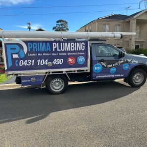 A truck with a prominently displayed service advertisement for plumbing. The vehicle features a blue and white colour scheme with the text 'PRIMA PLUMBING' and contact details. It is parked on a residential street, showcasing a suburban environment with houses in the background. The truck is equipped with plumbing tools visible on the roof rack, indicating its readiness for emergency plumbing services.